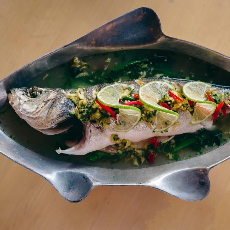 Steamed whole fish with lime slices, red chilies, and herbs in a fish-shaped serving dish.