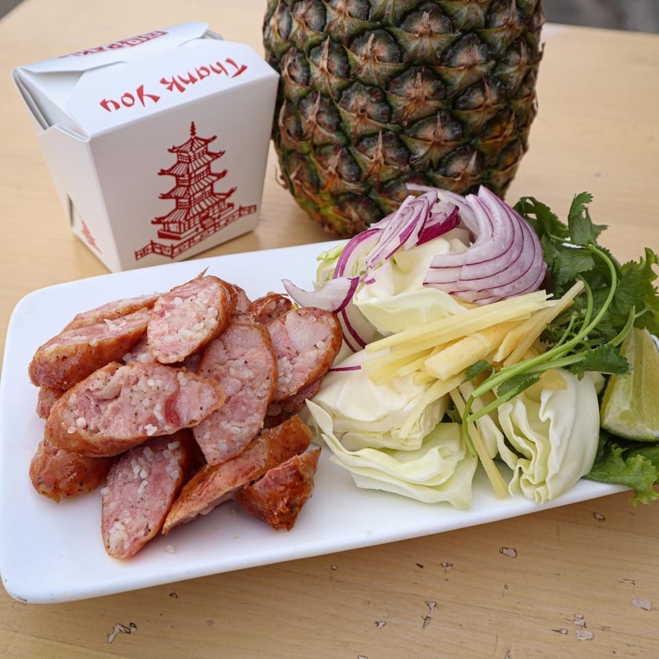 Thai sausage slices with cabbage, red onion, ginger, cilantro, and lime, next to a pineapple and takeout box.