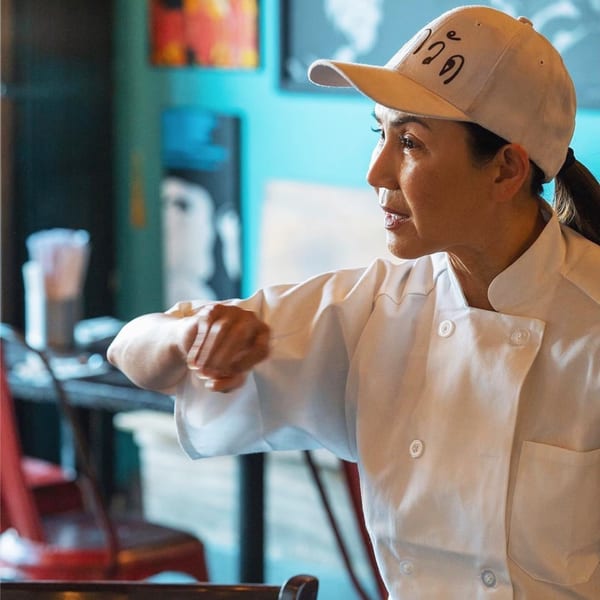 Chef in a white uniform and cap, with a ponytail, looking intently to the right.