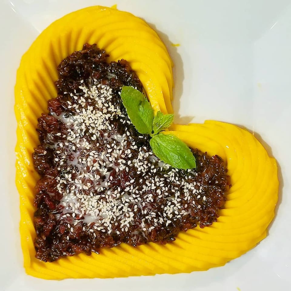 Heart-shaped mango slices surround dark sticky rice with sesame seeds and a mint sprig.