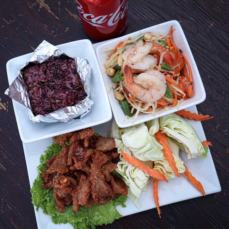 Thai meal with shrimp papaya salad, fried pork, purple sticky rice, cabbage, carrots, and a Coke.