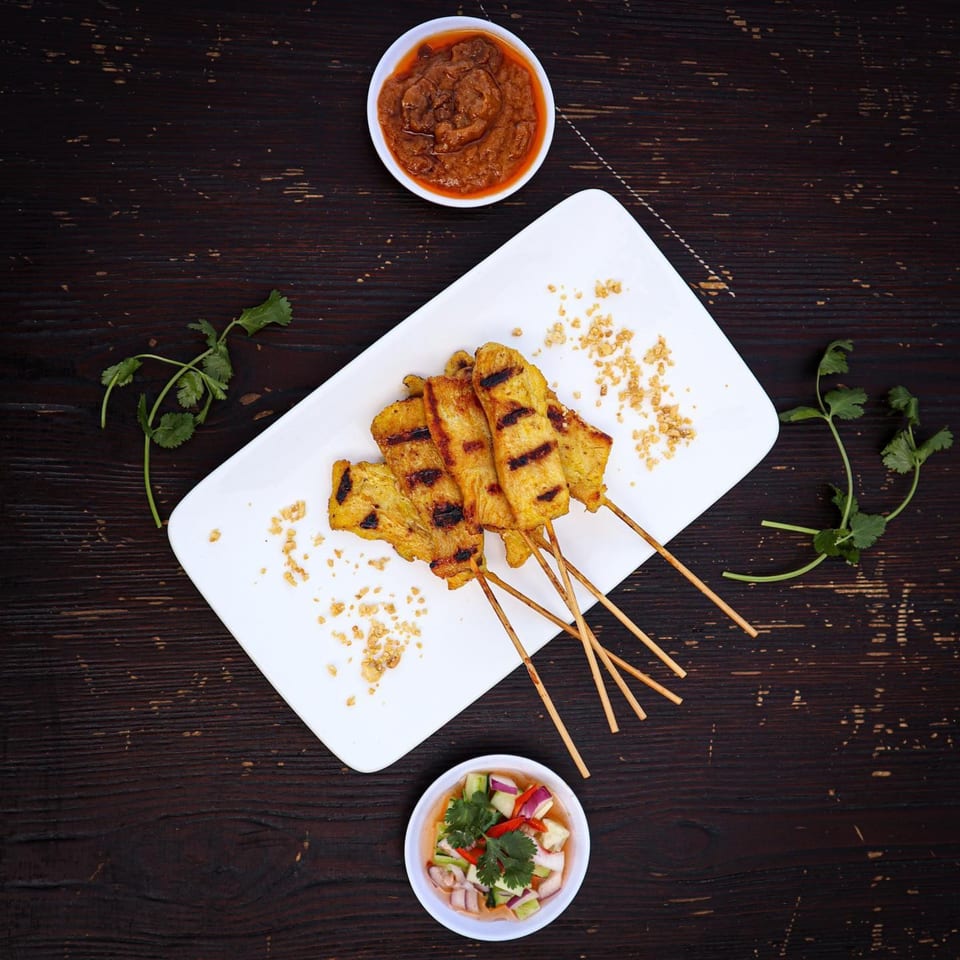 Grilled chicken satay skewers with peanut sauce and cucumber relish on a dark wooden table.