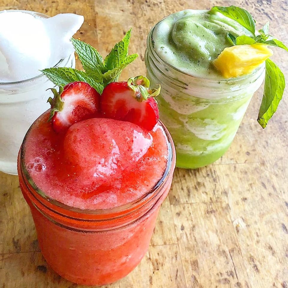 Two vibrant smoothies, one red with strawberries and mint, one green with pineapple and basil, on a wooden surface.